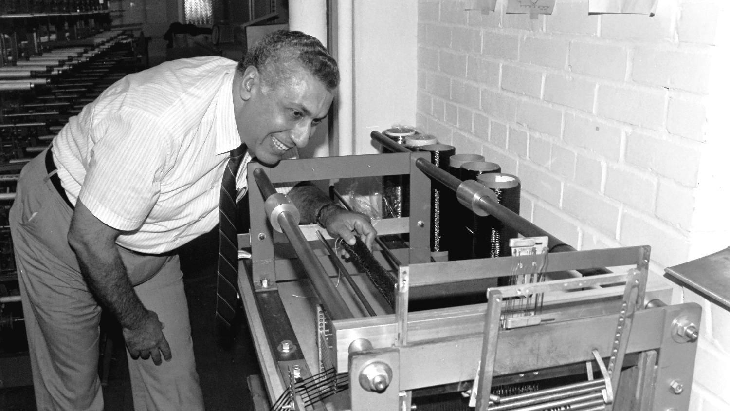 Mansour Mohamed smiles as he reaches his hand into a weaving machine in an NC State laboratory against a white brick wall. He wears dress pants and a white short-sleeved button up shirt with a dark, striped tie. Behind him is more machinery in black and white.