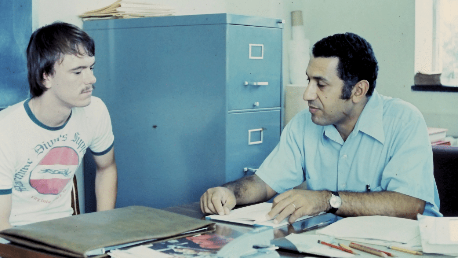 A color film image of Mansour Mohamed teaching a student. Mohamed is seated and wearing a blue short-sleeved button up shirt, and the student at the left of the frame wears a white t-shirt. They both look down at the paper Mohamed holds on the desk, which is cluttered with other papers and folders. They are seated in front of a light blue filing cabinet.