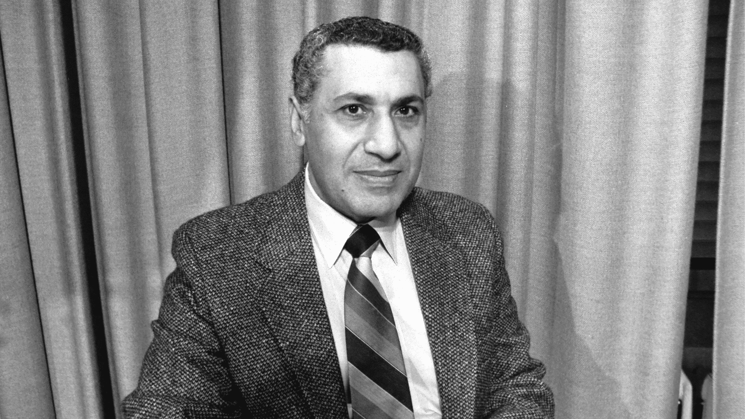 A black and white portrait of Mansour Mohamed. He is looking straight at the camera and he fills the middle of the frame. He wears a dark suit and a white shirt with a striped tie. His hair is closely cropped to his head. He is seated in front of a lightly colored curtain.