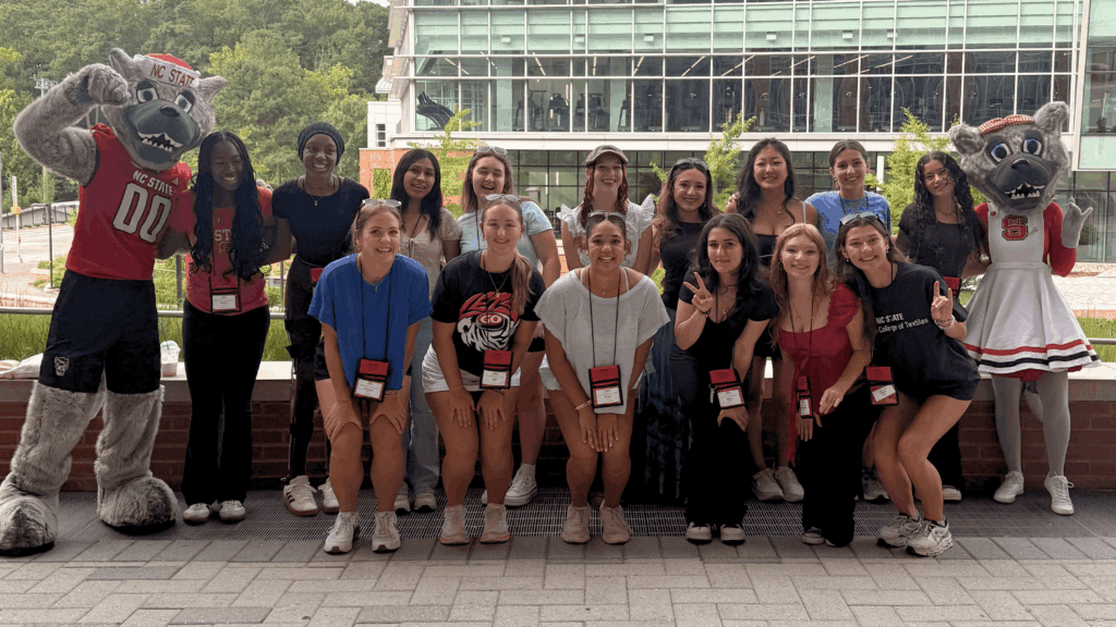A group of STEP students pose for a photo with Mr. and Ms. Wuf. They are lined up in two rows with the mascots at the left and right of the frame, with Carmichael Gym in the daylight behind them.