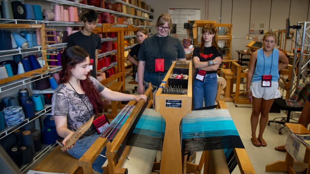 A high school student with long dark hair uses a hand loom. She holds the boat of the loom which is threaded with several colors of blue thread. Behind her is a colorful wall of spools. More looms stand in the background and other students stand and watch the demonstration.