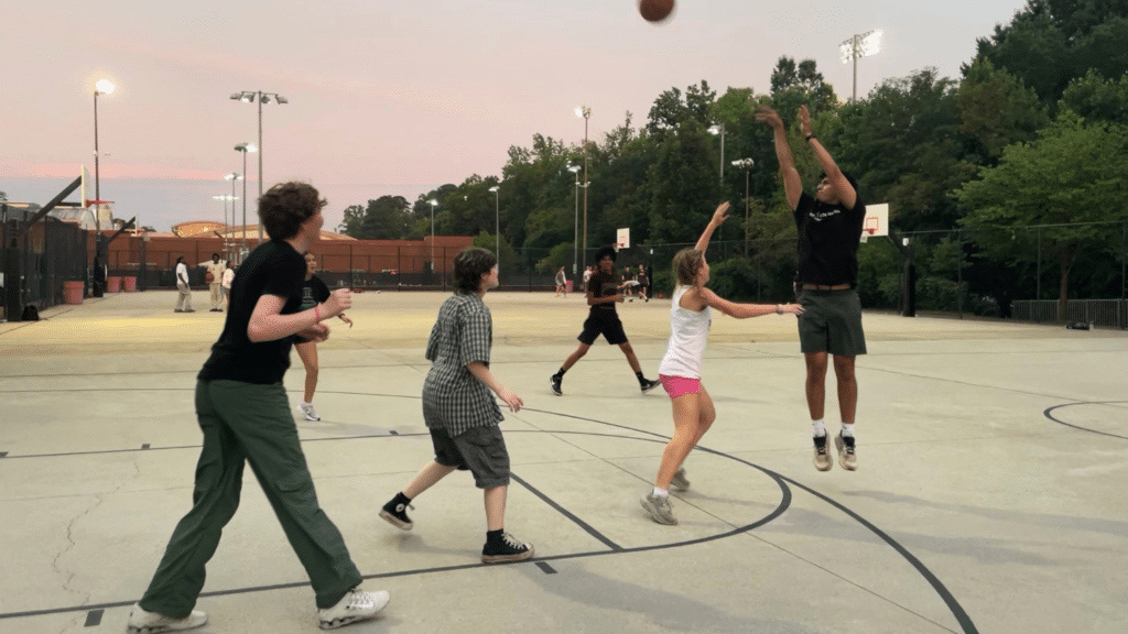 STEP students play basketball on the courts outside of Carmichael Gym. One is leaping and shooting the ball over the head of three others, who are in defensive positions. They are playing on a concrete court during sunset with a line of trees behind them.