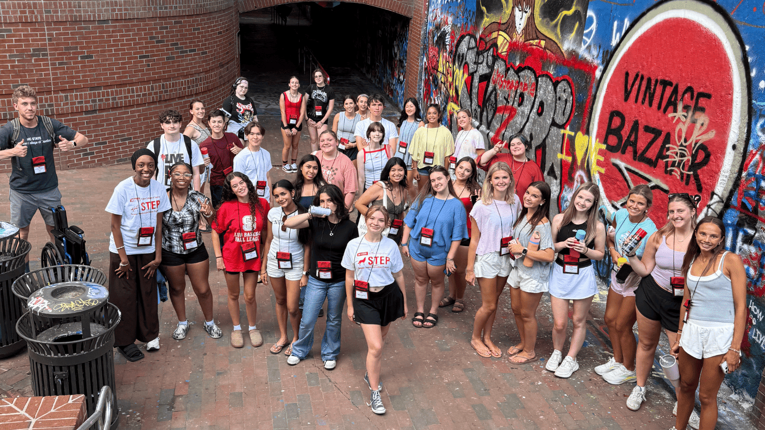 STEP students pose for a photo in front of the Free Expression Tunnel on Main Campus. The large wall near the entrance is covered in colorful paint and leads into the tunnel behind them. There a two dozen students wearing lanyards and summer clothing smiling at the camera.