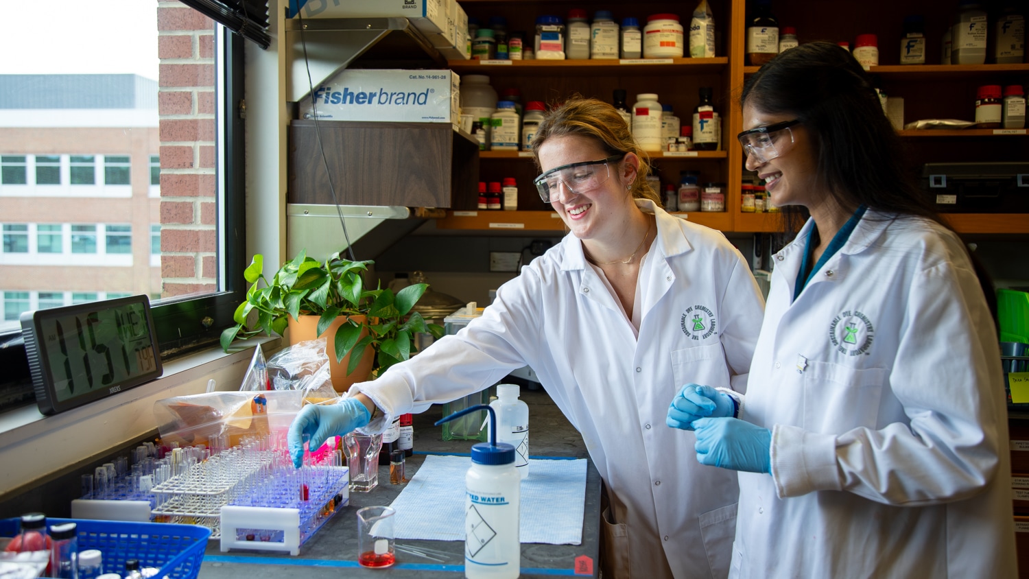 Regan Stefanoni and Krusha Patel work in a laboratory, wearing long white lab coats. Bathed in natural light from a window on the left, they both face a counter with racks of small beakers and other lab equipment. They smile as Regan reaches for one of the beakers.