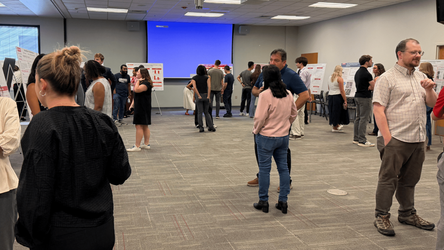 A wide shot of people gathered in a conference room for the TECS REU Research Symposium. Posters line the circumference of the room and students present their work to onlookers, who populate the foreground and background.