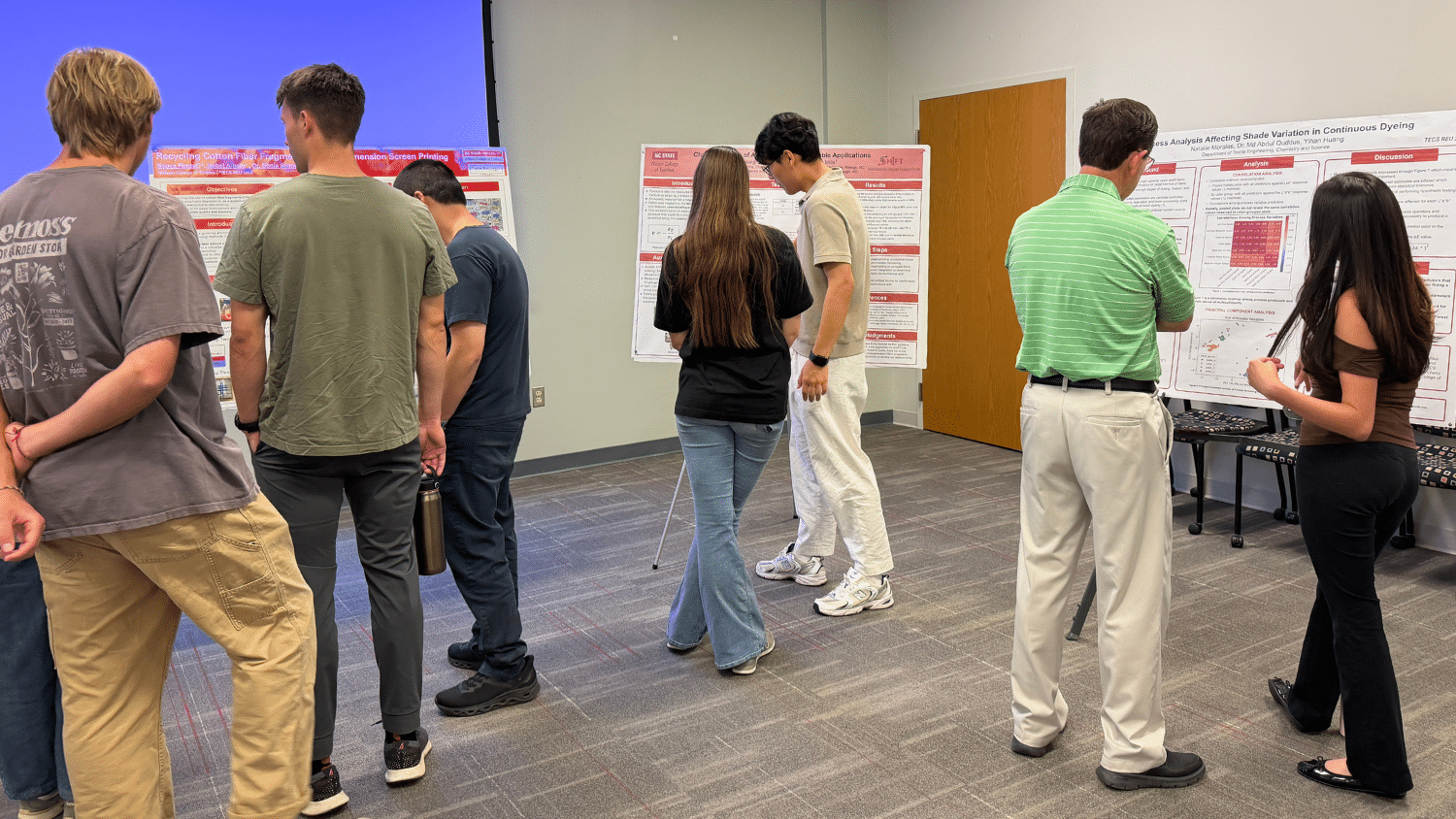 People gathered in a conference room for the TECS REU Research Symposium. Three posters in the corner of the room are being read by onlookers.