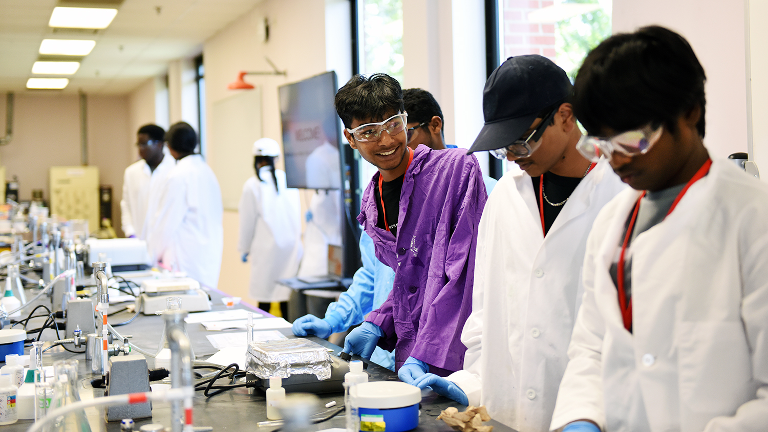 High school students wearing lab coats participate in a lab exercise at TexTEC camp. They are in a lab and facing a counter with measuring instruments and sinks. One student wearing a purple lab coat and goggles leans against the counter and smiles at his lab partner.