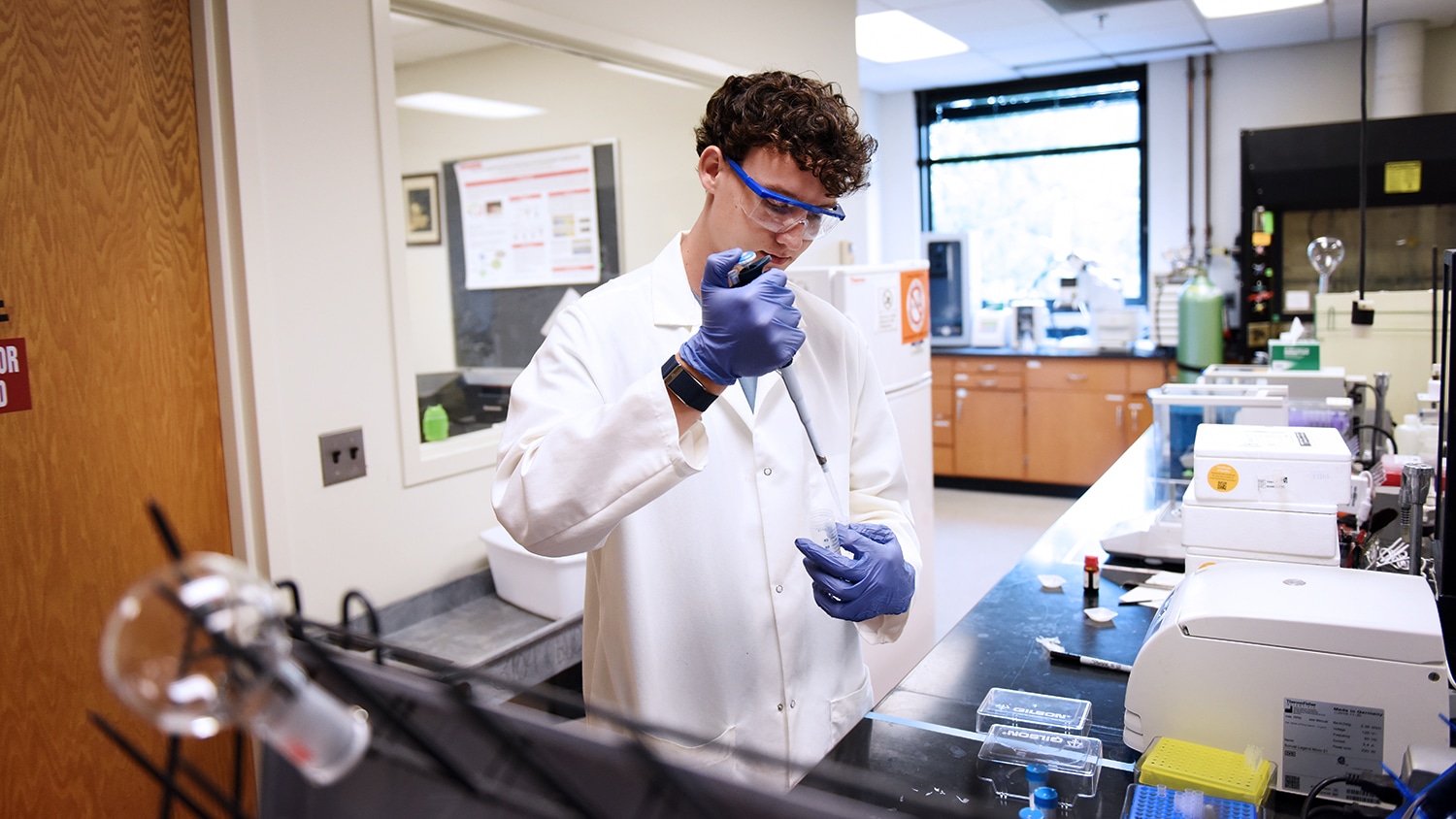 Connery Bunn uses a measuring instrument in his research lab. He wears a white lab coat and goggles. Around him is more lab equipment and a behind him is a bright window, flooding the room with natural light.