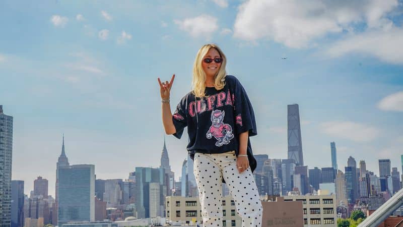 Katie Watson holding up the Wolfpack Symbol as she stands on a rooftop in New York City with the skyline behind her