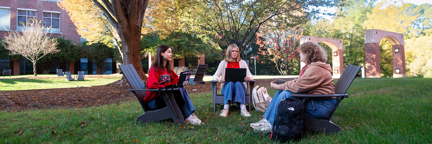 Two students sit outside on steps, smiling and making the "wolfs up" sign. One wears a red NC&#160;State shirt and the other a black top with an NC&#160;State logo. Greenery and part of the Wilson College of Textiles are in the background.