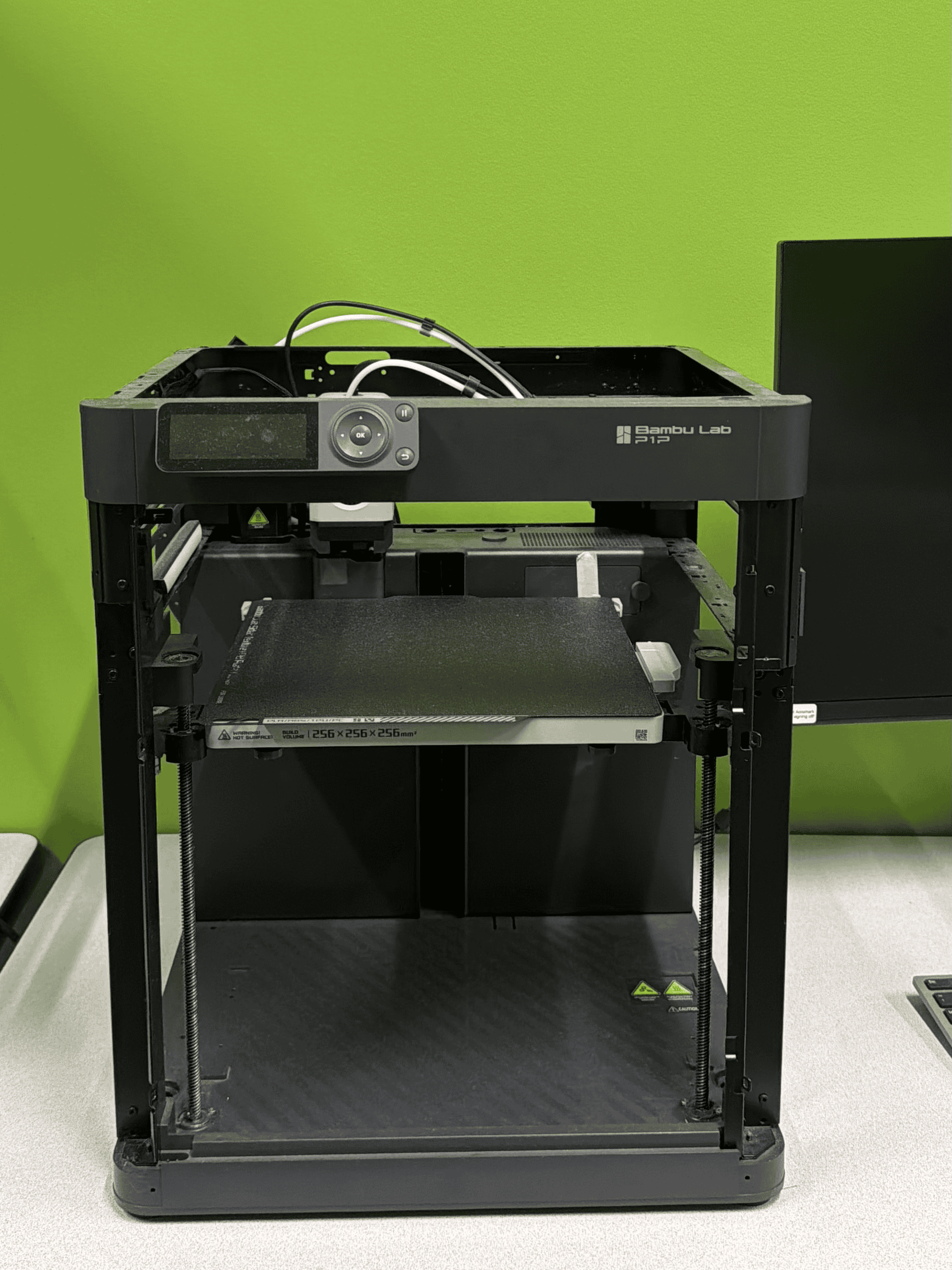 A Bambu Lab P1P desktop 3D printer, part of the Bambu Lab P1 series, equipped with an Automatic Material System (AMS) mounted on top. The AMS contains four filament spools (black, red, white, and green), and two completed 