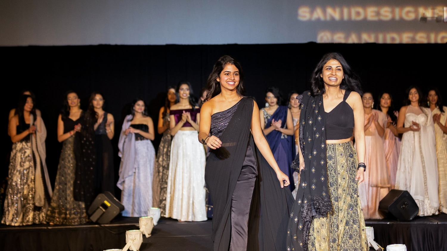 Ritika (left) and Nikie (right) Shamdasani walk down a runway. Models are visible in the background. 